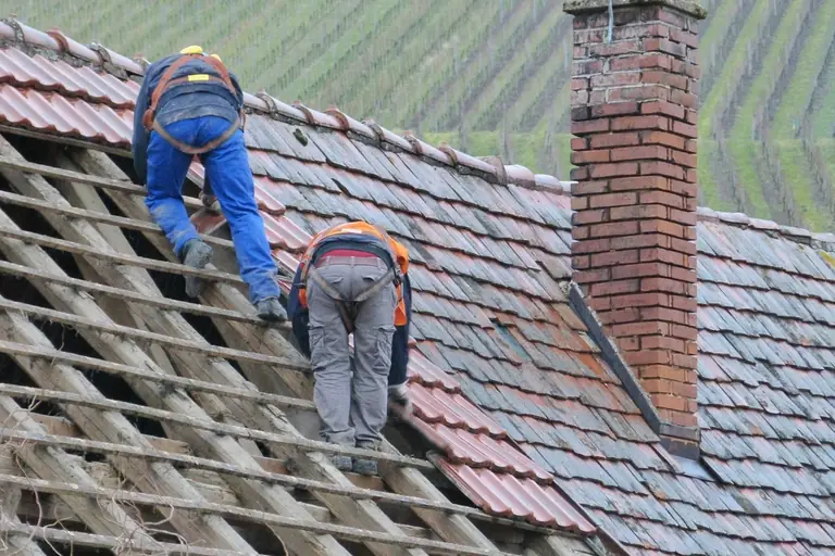 Two roofers on a tile roof beside a chimney